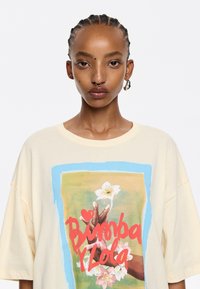 Kremowy, oversize'owy T-shirt z kolorową grafiką przedstawiającą dłonie i kwiaty. Gładka bawełniana tekstura, krótki rękaw i okrągły dekolt.