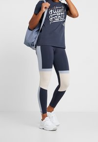 Námořnicky modrý a světle šedý sportovní outfit zahrnující tričko s potiskem a legíny s barevnými bloky. Bílá teniska doplňuje vzhled.