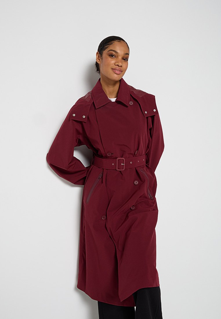 Bordeaux trenchcoat met een ceintuur in de taille, dubbele knopen, schouderepaulets en twee zijzakken, weergegeven tegen een neutrale achtergrond.