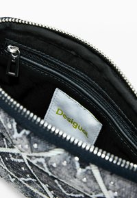 Szürke, texturált anyagból készült tasak ezüst cipzárral, szegecsekkel díszítve és egy sárga "Desigual" feliratos címkével. A belső rész fekete.