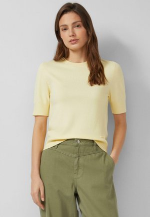 Woman with long brown hair wearing a light yellow short-sleeve sweater and olive green high-waisted pants, standing with one hand in pocket.