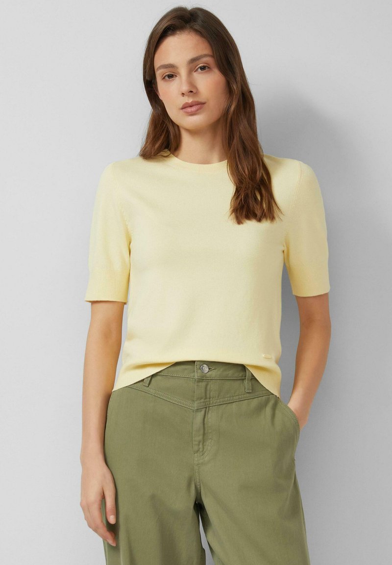 Vrouw met lang bruin haar, gekleed in een lichtgeel truitje met korte mouwen en olijfgroene broek met hoge taille, staand met één hand in de zak.