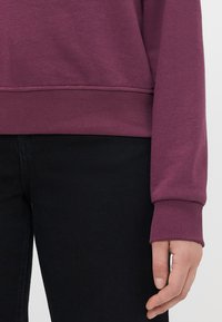 Persona che indossa una felpa viola scuro con polsini e orlo a costine, e pantaloni neri, con la mano rilassata al suo fianco.