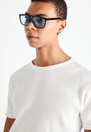 Crne sunčane naočale s plavim staklima, pravokutnog oblika, nošene uz teksturiranu bijelu pletenu majicu s okruglim izrezom i kratkim rukavima.