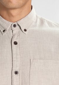 Jasnobeżowa koszula na guziki z teksturowanej tkaniny, z ko�łnierzykiem, brązowymi guzikami i pojedynczą kieszenią na piersi.