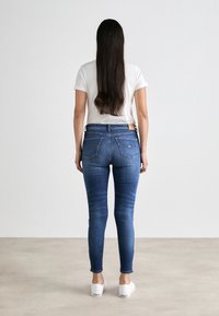 Jeans ajustados en denim azul oscuro, con una cintura de altura media, cinco bolsillos y un desvanecimiento sutil, combinados con una camiseta casual blanca y zapatos blancos.