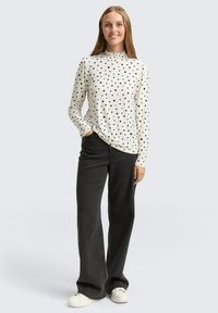 Langärmliges weißes Oberteil mit schwarzen Punkten, das einen gerafften hohen Kragen hat, kombiniert mit weit geschnittenen schwarzen Jeans und weißen Sneakern.