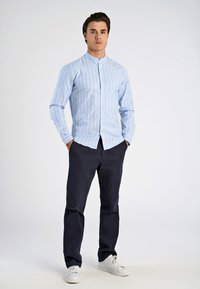 Camisa de rayas azul claro con botones y cuello mao, combinada con pantalones de un azul marino oscuro y zapatillas blancas. La camisa tiene rayas blancas verticales.