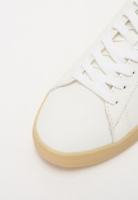 White leather sneaker with a rounded toe, flat white laces, and a rubber gum sole. Seam detailing along the side adds texture.