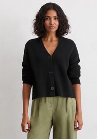 Schwarze Strickjacke mit V-Ausschnitt, drei Knöpfen auf der Vorderseite und leicht verkürzten Ärmeln. Getragen über olivgrünen Hosen.