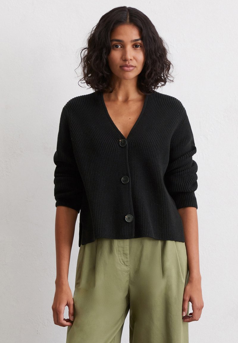 Schwarze Strickjacke mit V-Ausschnitt, drei Knöpfen auf der Vorderseite und leicht verkürzten Ärmeln. Getragen über olivgrünen Hosen.