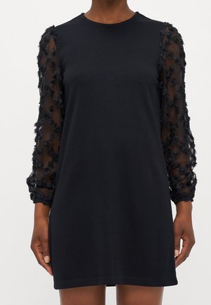 Czarna sukienka z przezroczystymi długimi rękawami ozdobionymi aplikacjami 3D w kształcie kwiatów, z okrągłym dekoltem, noszona przez osobę o średnio-ciemnej karnacji.