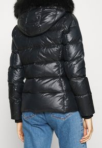 Person som bär en svart kviltad puffjacka med huva med pälsdetalj och blå denimjeans, sedd bakifrån.