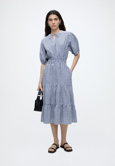 Femme portant une robe midi vichy bleue et blanche avec des manches bouffantes, tenant un sac à main noir et portant des sandales noires, debout devant un fond blanc.