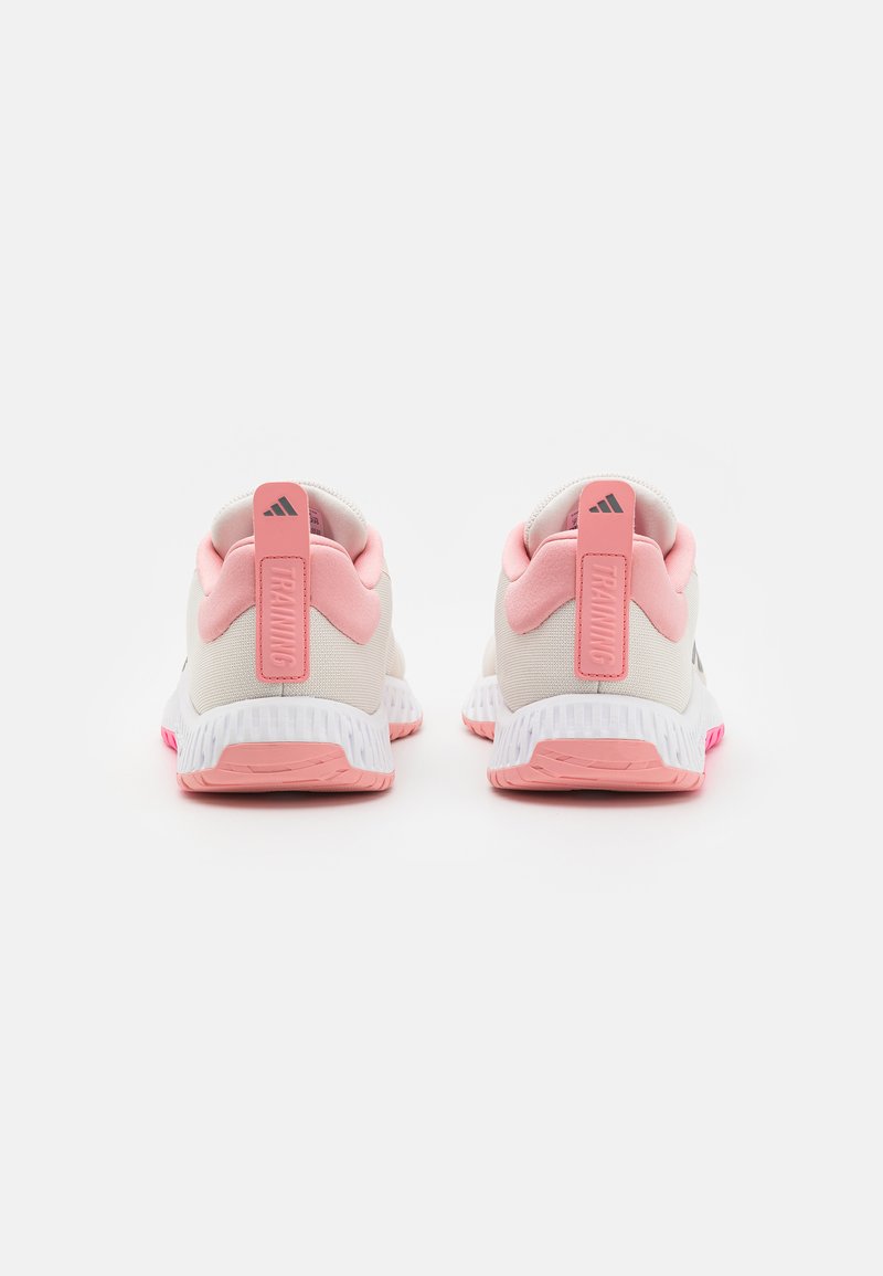 Calzado con una parte superior blanca, detalles en rosa y una suela blanca estriada. Presenta un prominente tirador en el talón rosa etiquetado como "ENTRENAMIENTO."
