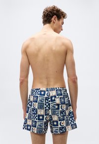 Νεαρός άνδρας χωρίς μπλούζα, εμφανιζόμενος από πίσω, φοράει μπλε και λευκό μαγιό με σχέδια ήλιου, φεγγαριού και φοίνικα.