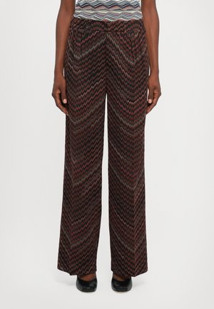 Persoană îmbrăcată în pantaloni cu picioare largi, cu un model zigzag multicolor și pantofi negri, stând în fața unui fundal simplu de culoare deschisă.