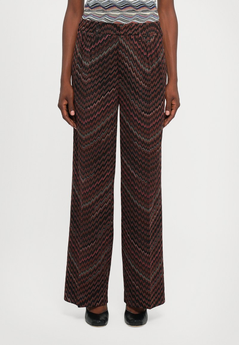 Persona che indossa pantaloni a gamba larga con motivo zigzag multicolore e scarpe nere, in piedi su uno sfondo chiaro e uniforme.