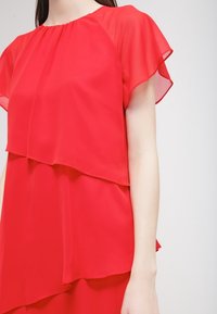 Haut en chiffon rouge avec un design superposé, manches courtes et col rond. Le tissu semble léger et semi-transparent.