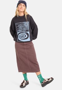 Svart oversized genser med blå grafikktrykk, brun midi-skjørt, grønn mønstrete sokker og ku-mønstrede sko. Lue på hodet.