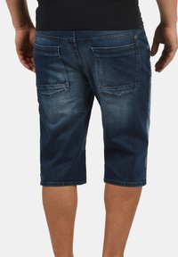 Tummansinisiä farkkushortsit. Keski-reiden pituus, joissa on kaksi takataskua, ommellut yksityiskohdat ja hieman haalistunut pinta.