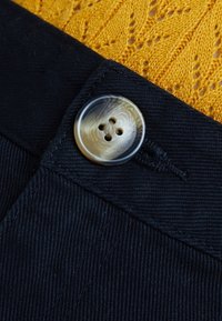 Tessuto nero con un grande bottone rotondo marmorizzato a quattro fori. Un maglione texture color giallo senape è parzialmente visibile sullo sfondo.