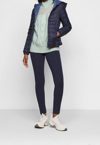 Daunenjacke mit navy Außenfarbe und hellblauem Innenfutter, kombiniert mit einem hellgrünen Zopfmuster-Pullover, navy Leggings und weißen Sportschuhen.