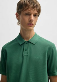 Grön polo skjorta med texturerad yta, två knappar och en diskret logotyp på bröstet. Har en nedvikt krage och korta ärmar.