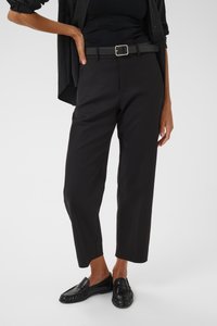 Persona che indossa pantaloni corti neri con una cintura di pelle nera e mocassini neri, in piedi davanti a uno sfondo semplice.