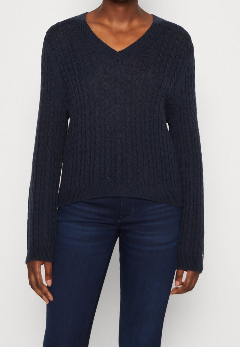 Personne portant un pull en maille torsadée bleu marine en V et un jean ajusté bleu foncé sur fond blanc uni.