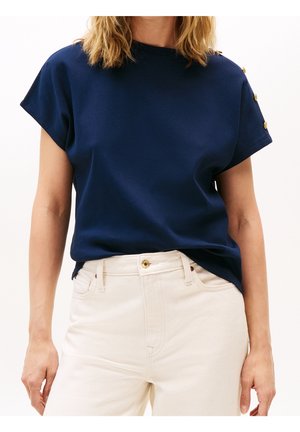 Femme portant un haut bleu marine à manches courtes avec des boutons dorés sur l'épaule et un pantalon blanc taille haute, debout sur fond blanc.