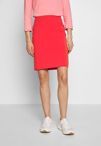 Gonna rossa aderente realizzata in materiale elastico, lunghezza al ginocchio, con una texture liscia. Indossata con scarpe da ginnastica leggere e una maglia rosa a maniche lunghe.