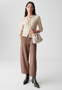 Beige gerippter Cardigan mit Knöpfen, braune hochtaillierte weit geschnittene Hose und elegante schwarze spitz zulaufende Stiefeletten; trägt eine hellgraue Zugtasche.