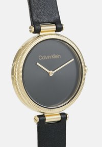 Calvin Klein laikrodis su juoda odine apyranke, apvaliu pilku ciferblatu, auksinės spalvos rėmeliu su grioveliais ir paprastomis auksinėmis rodyklėmis.