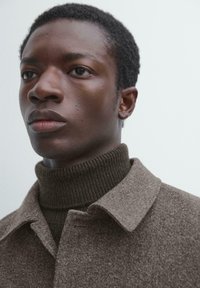 Brun uldfrakke med krave, lagdelt over en mørk, ribbet rullekrave. Hudtonen er mellem, håret er kort og struktureret.