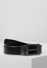 Ceinture en cuir noir avec une texture lisse, présentant une boucle en métal argenté rectangulaire embossée avec "Porsche Design." Design classique.