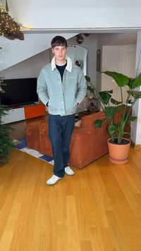 Quilted light blue jacket with cream collar, black shirt, dark jeans, and white sneakers. Indoor setting with plant and orange-accented furniture.