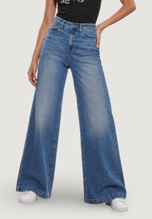 Donna che indossa jeans blu a vita alta con gambe larghe abbinati a sneaker bianche e un top nero senza maniche, posando con una mano sui fianchi.