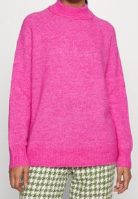 Fuchsia stickad tröja med hög krage, ledig passform, ribbade mudd och fåll; kombinerad med gröna och vita byxor i hundtandsmönster.