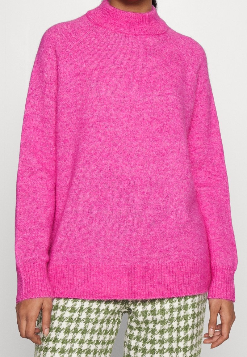 Fuchsia stickad tröja med hög krage, ledig passform, ribbade mudd och fåll; kombinerad med gröna och vita byxor i hundtandsmönster.