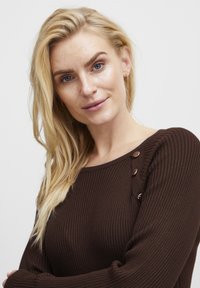 Maglione marrone a coste con scollatura rotonda e dettagli in bottone sulla spalla. La texture morbida e il design aderente ne esaltano la struttura.