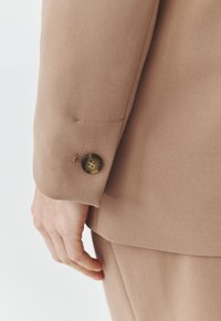 Gros plan d'une manche de costume beige avec un petit bouton marron et une main reposant à côté sur un fond uni.