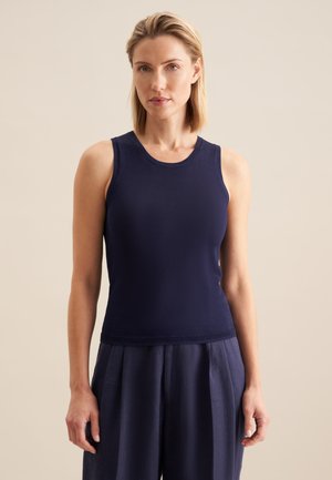 Vrouw met kort blond haar, die een mouwloos marineblauw topje en losse marineblauwe broek draagt, staand tegen een lichte achtergrond.
