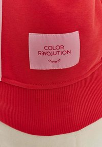 Červená mikina vyrobená z materiálu s ribbovaným lemem. Na ní je růžová nášivka s nápisem "COLOR R3VOLUTION" a jednoduchou grafikou řas.