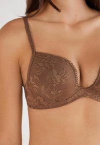 Close-up van een vrouw die een bruine bloemenkant bh met verstelbare banden draagt tegen een effen achtergrond.
