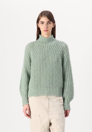 Mintgroene gebreide trui met een hoge nek, geribbelde textuur, losse pasvorm en lange mouwen. Draagt over crèmekleurige broek.