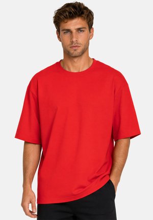 Jonge man met kort bruin haar draagt een los rood T-shirt en een zwarte broek, staand tegen een effen lichtgrijze achtergrond, kijkend naar voren.
