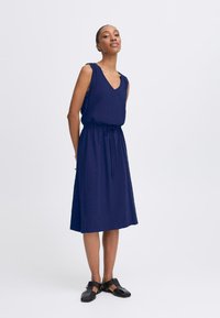 Navy midi dress with a V-neck, sleeveless design, elastic waist with drawstring, made from lightweight fabric, paired with black sandals.