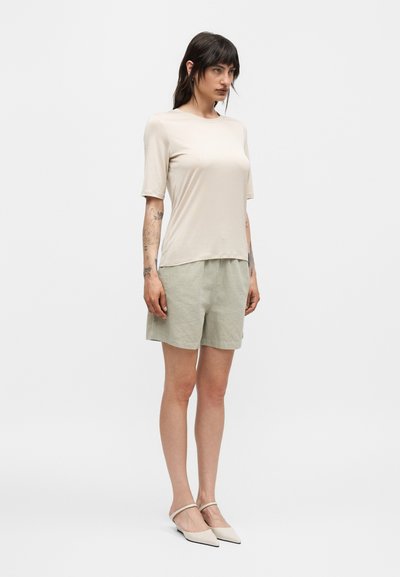 Γυναίκα με σκουρόχρωμα μαλλιά που φοράει μπεζ τοπ με κοντά μανίκια, ανοιχτοπράσινα σορτς και μπεζ μυτερά τακούνια, στέκεται μπροστά σε ένα απλό φόντο.