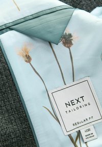 Svijetloplava tkanina s cvjetnim uzorkom i tirkiznim obrubom, sašivena oznaka glasi "NEXT TAILORING REGULAR FIT 40R," na teksturiranoj tamnosivoj pozadini.